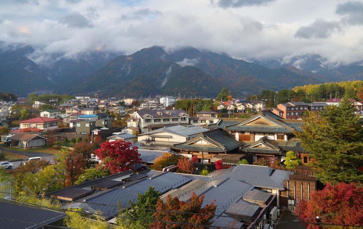 紅葉した木々のあいだに瓦屋根や金属屋根の住宅が並び、その奥に雲のかかった山並みが見える