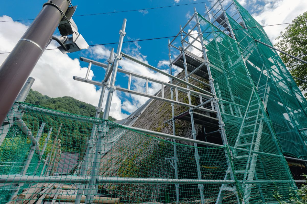 建物のまわりに銀色の足場が組まれ、緑色の防護ネットが張られている。下から見上げた構図